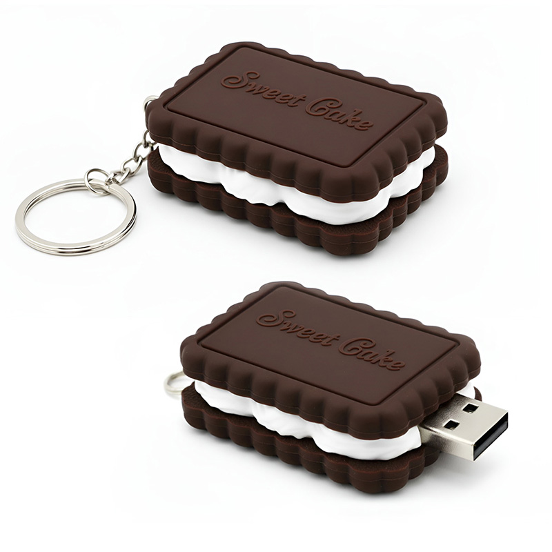 A 3D USB pendrive shaped like a cookie – two dark brown biscuits with white cream between them, the top biscuit embossed with the 3D inscription “Sweet Cake.” Features a metal keyring and realistic cookie texture. No cap. Designed by the Polish manufacturer DECARD