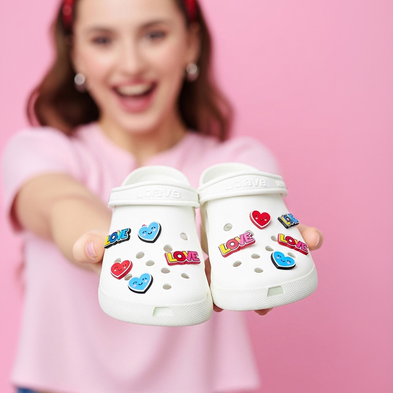 A young woman holding white Crocs decorated with colorful charms in the shape of hearts and the word LOVE, made from raised resin. Manufactured in Poland by DECARD